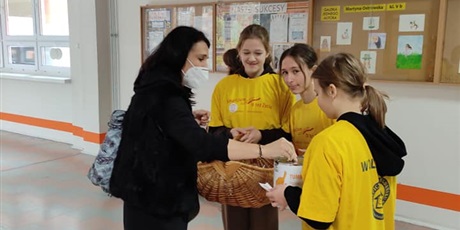 Powiększ grafikę: Dziewczyny ubrane w żółte koszulki wolontariatu trzymają koszyk. Nauczycielka wrzuca pieniądze do puszki ze zbiórką.