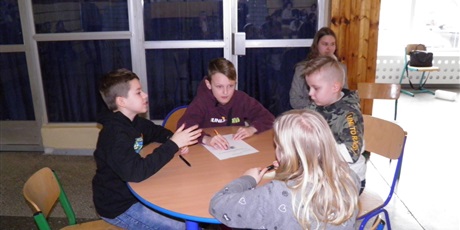 Powiększ grafikę: Czworo dzieci, 3 chłopców i dziewczynka. Siedzą przy okrągłym stoliku na którym leży kartka. 