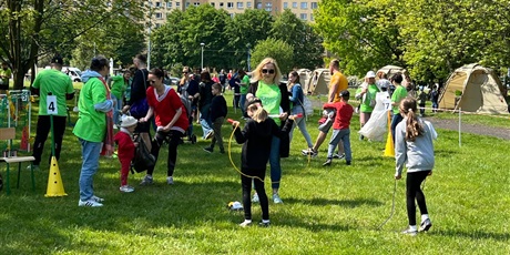 Powiększ grafikę: Podczas festynu, na trawie widoczne są bawiące się dzieci birące udział w różnych konkurencjach sportowych. Opiekują się nimi nauczyciele.