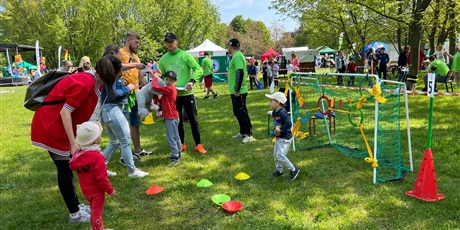 Powiększ grafikę: Podczas festynu, na trawie widoczne są bawiące się dzieci birące udział w różnych konkurencjach sportowych. Opiekują się nimi nauczyciele.