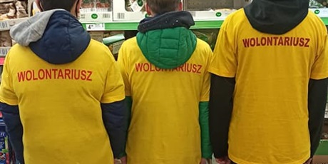 Powiększ grafikę: Trzej chłopcy ubrani w żółte koszulki. stoją w sklepie, odwróceni tyłem pokazując napis na koszulce "Wolontariusz".