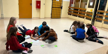 Powiększ grafikę: 2 grupy dzieci siedzące na podłodze i rozwiązujące zdania
