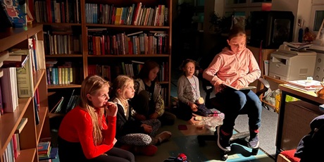 Powiększ grafikę: Uczennica czyta książkę w świetle lampki nocnej, wokół zasłuchane dzieci, w tle regały z książkami.