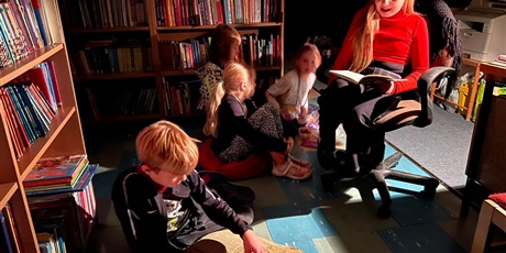 Powiększ grafikę: Uczennica czyta książkę w świetle lampki nocnej, wokół zasłuchane dzieci, w tle regały z książkami.